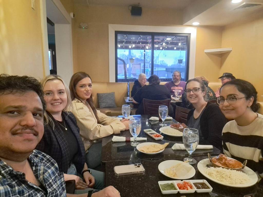 The image shows Lindsey and Farimah at dinner with students and the faculty host, Anoop Arunagiri.